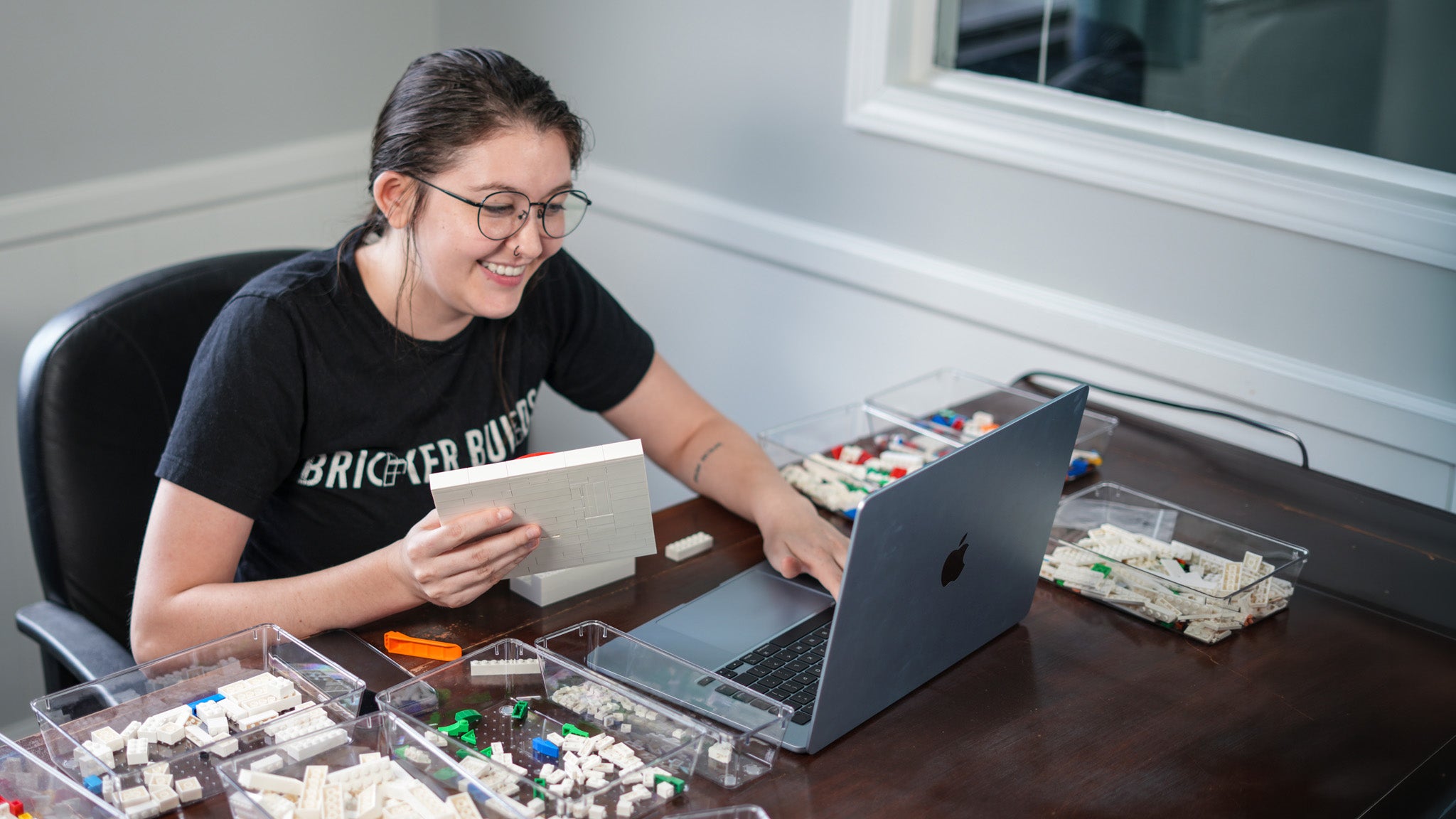 Una mujer con gafas y una camiseta negra con el gráfico de Bricker Builds está sentada en un escritorio de madera, sonriendo mientras sostiene un cuaderno blanco y usa una laptop plateada. Hay contenedores plásticos transparentes con bloques pequeños sobre el escritorio frente a ella.